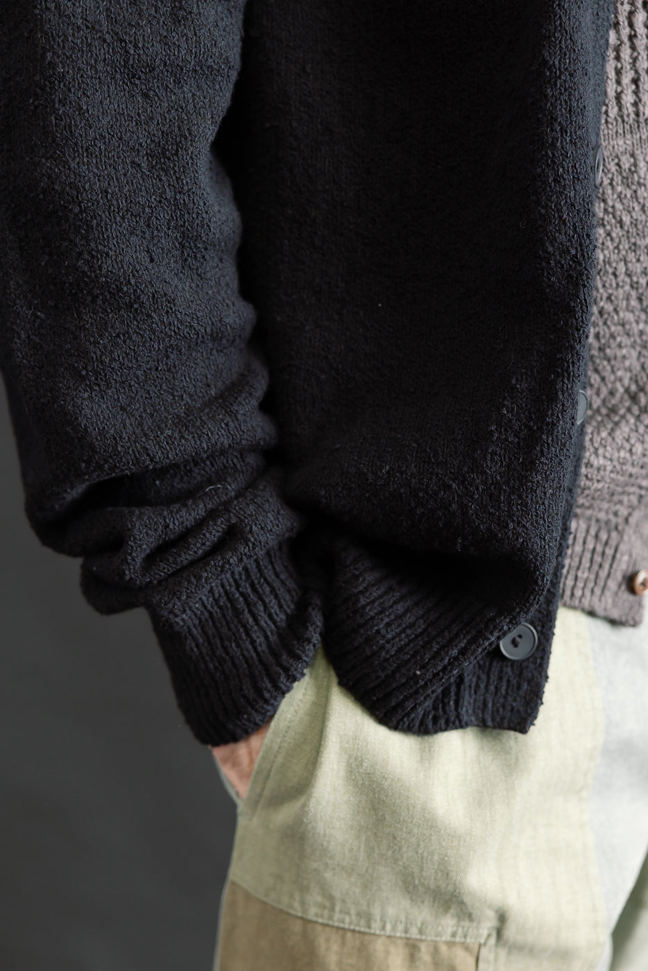 Close-up of a male model's lower right torso wearing a black boucle cardigan, showing textured fabric, ribbed cuff, and side front buttons. Model is 5'9