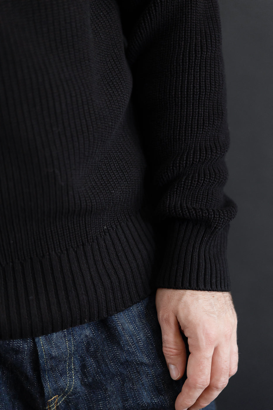 Close-up side view of a black ribbed knit sweater and dark selvedge denim on a male model.