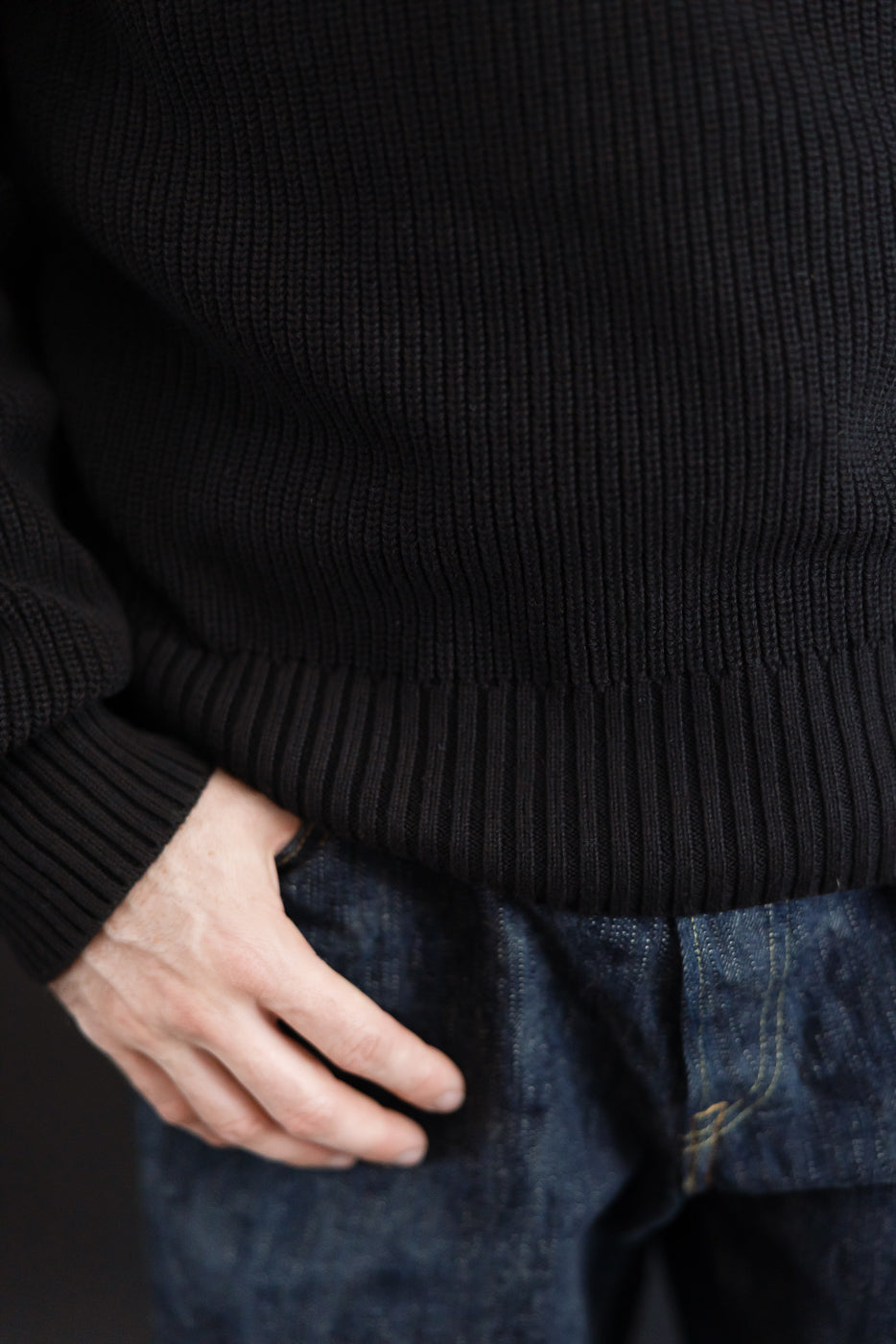 Close-up front view of a male model's black ribbed knit sweater hem and dark denim jeans.