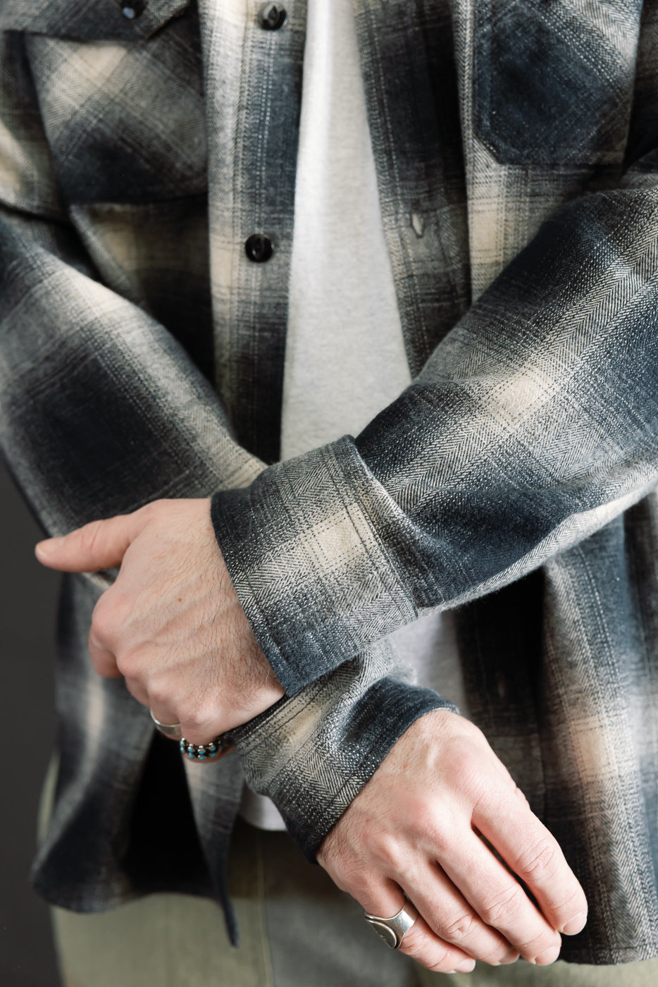 Close-up of a male model’s crossed arms wearing black ombre plaid flannel shirt, front details, showing buttoned cuffs and chest pockets. Model is 5'9