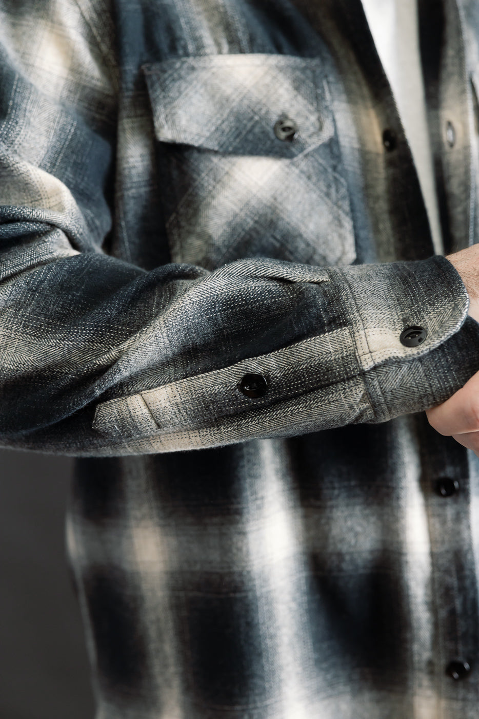 Close-up of black ombre plaid flannel shirt on male model, showing front left chest pocket, textured fabric, black buttons, and sleeve cuff detail. Model is 5'9