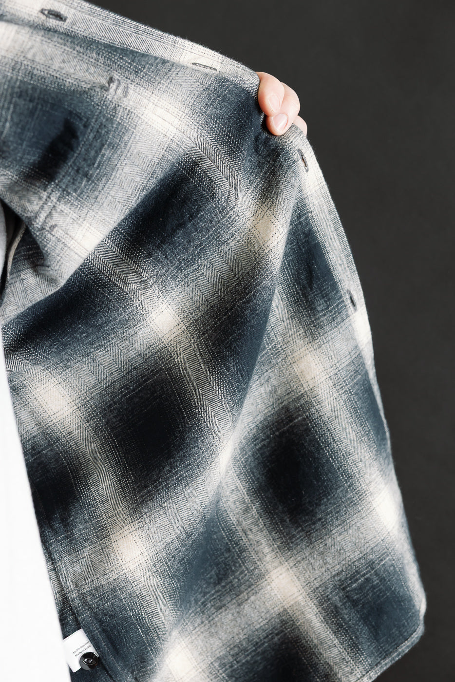 Close-up of male model holding up inside front of black ombre plaid flannel shirt, showing fabric texture and button detail.