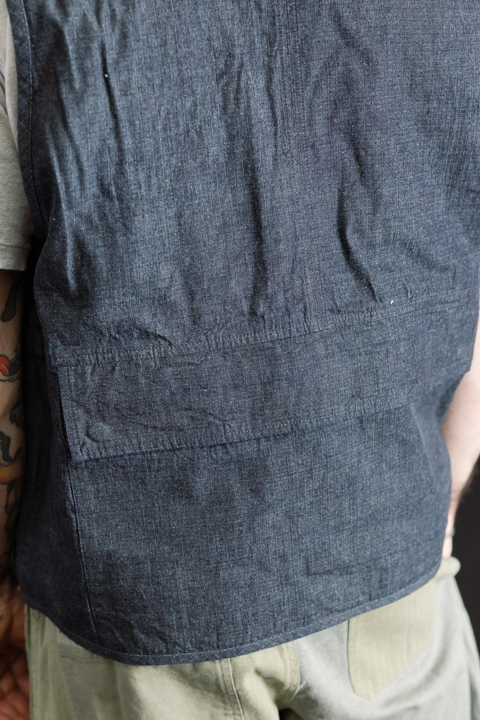Close-up of male model's back wearing indigo ripstop field vest, showing rear fabric and pocket detail. Model is 5'9