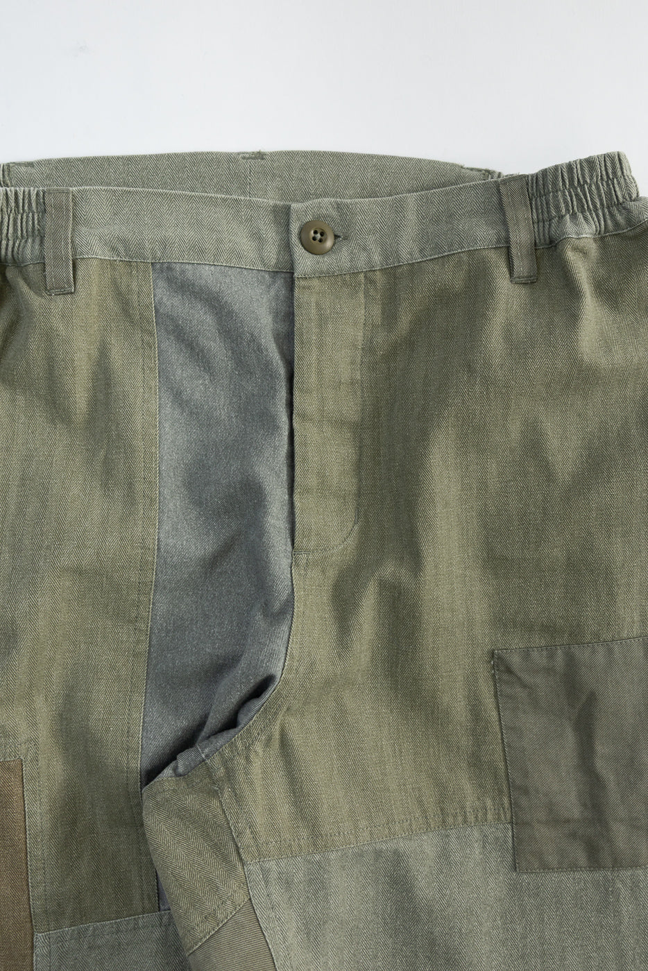 Close-up front view of olive patchwork pants, showing button, waistband, and textured fabric.