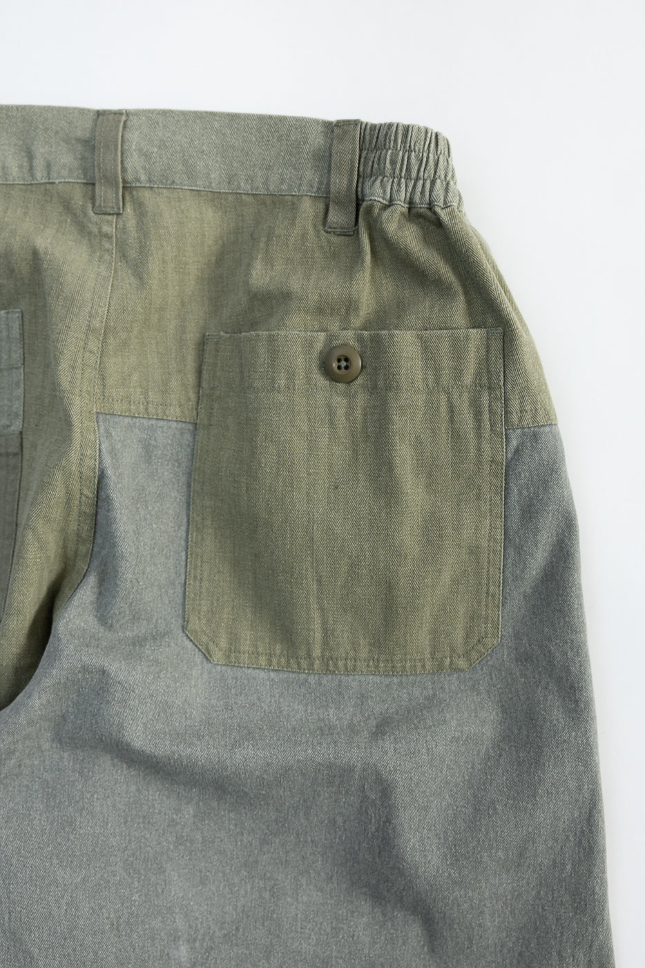 Close-up of back right patch pocket on olive patchwork pants, showing button and elastic waistband detail.