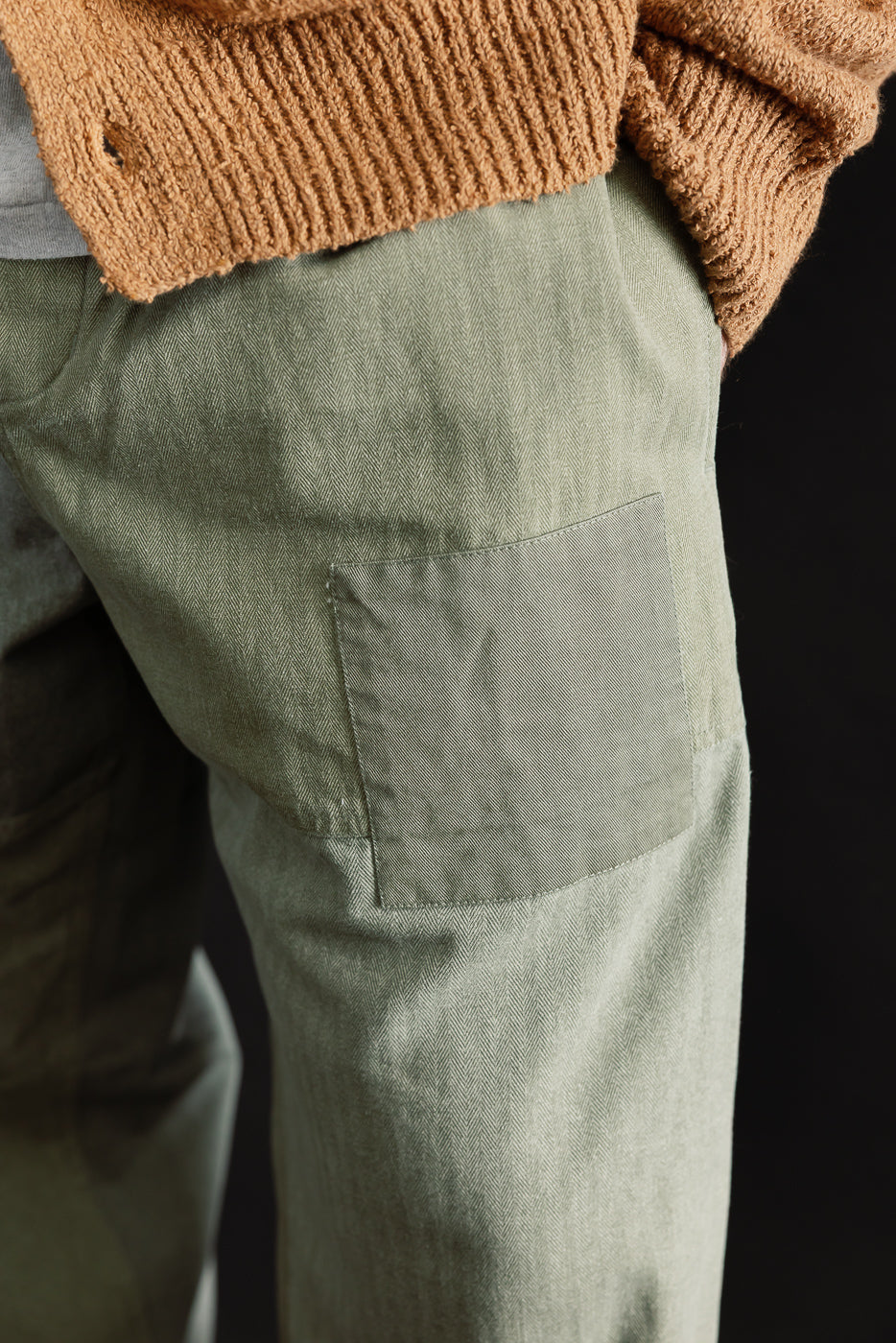 Close-up of male model's right hip wearing olive patchwork herringbone pants, visible pocket detail.