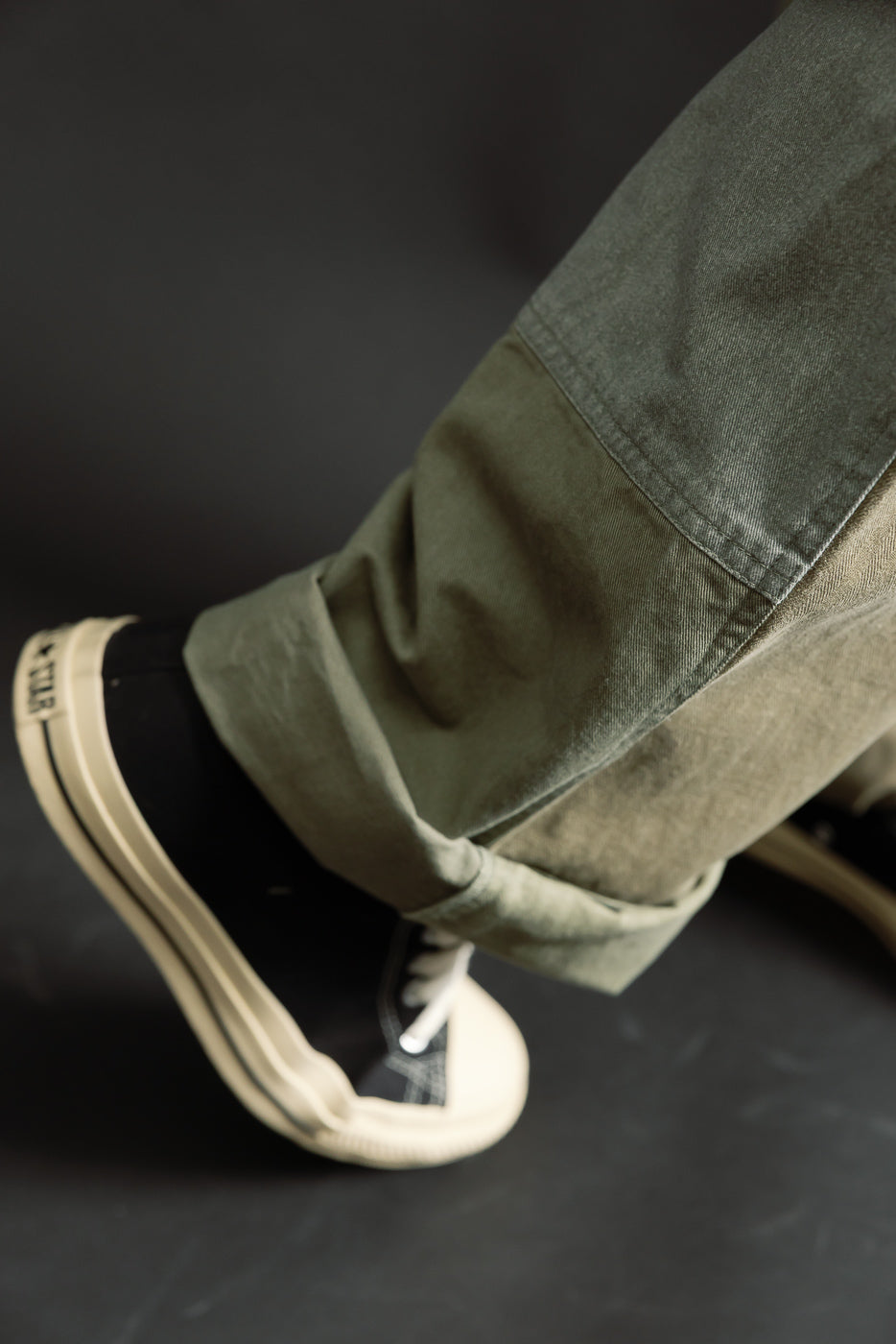 Close-up of male model's lower leg wearing olive patchwork pants, side view, cuffed hem detail, black sneakers visible. Model is 5'9