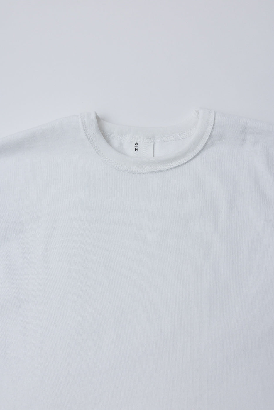 Close-up of the front upper chest and collar of a white heavyweight boxy t-shirt, showing ribbed crew neck and tag detail.