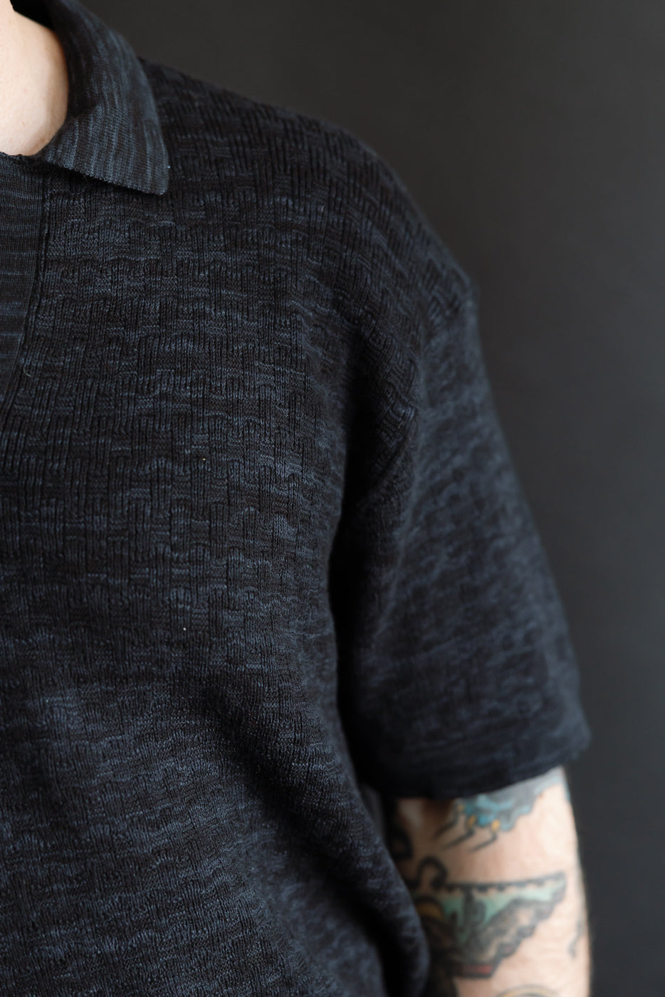 Close-up of male model’s front left side wearing knit polo in vintage black, highlighting textured boxweave fabric, collar, short sleeve, and athletic build. Model is 5'9