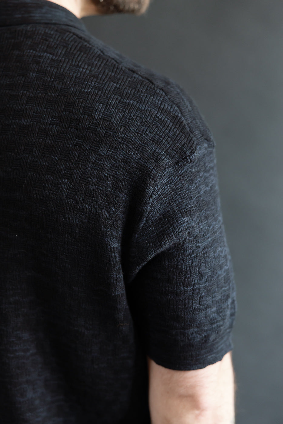 Close-up of male model's upper back and shoulder wearing black knit polo, showing textured boxweave fabric, short sleeve, rear side. Model is 5'9