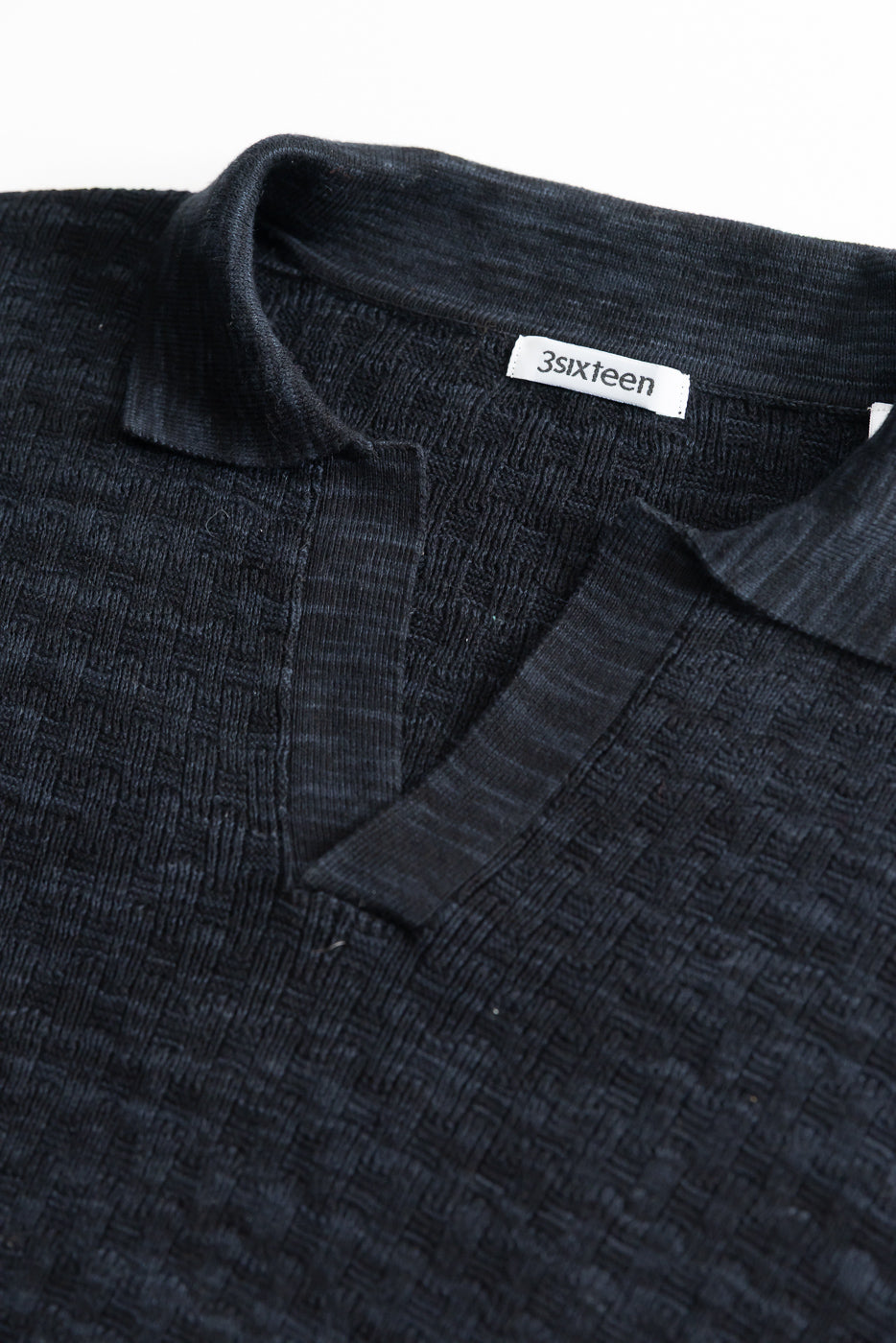 Close-up of front upper chest and collar of black knit polo, showing textured boxweave fabric, short collar, and placket detail.