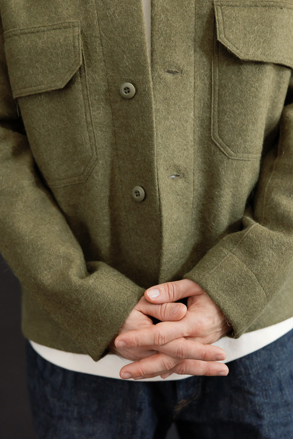 Close-up front view of a male model wearing an olive green wool shirt jacket with visible buttons and texture.