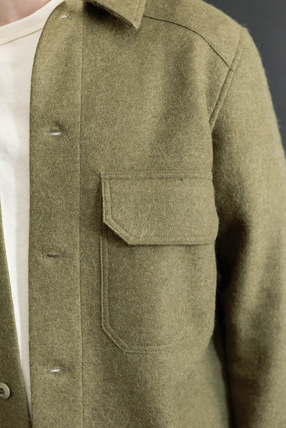 Close-up front view of an olive green wool shirt jacket showing chest pocket and fabric texture.