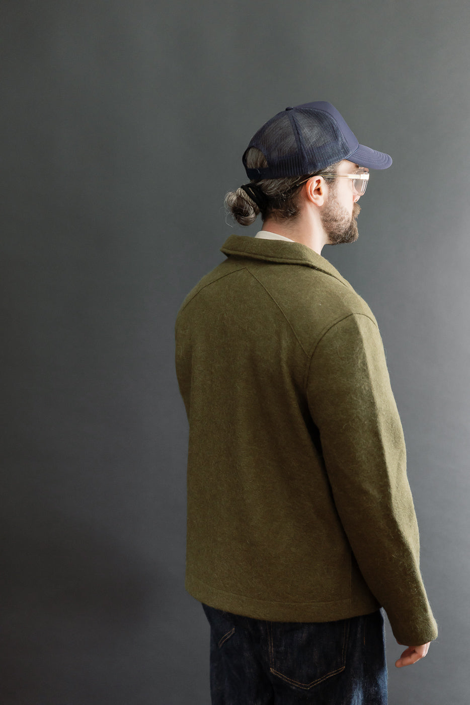 Back view of a male model wearing an olive green wool jacket and blue cap.
