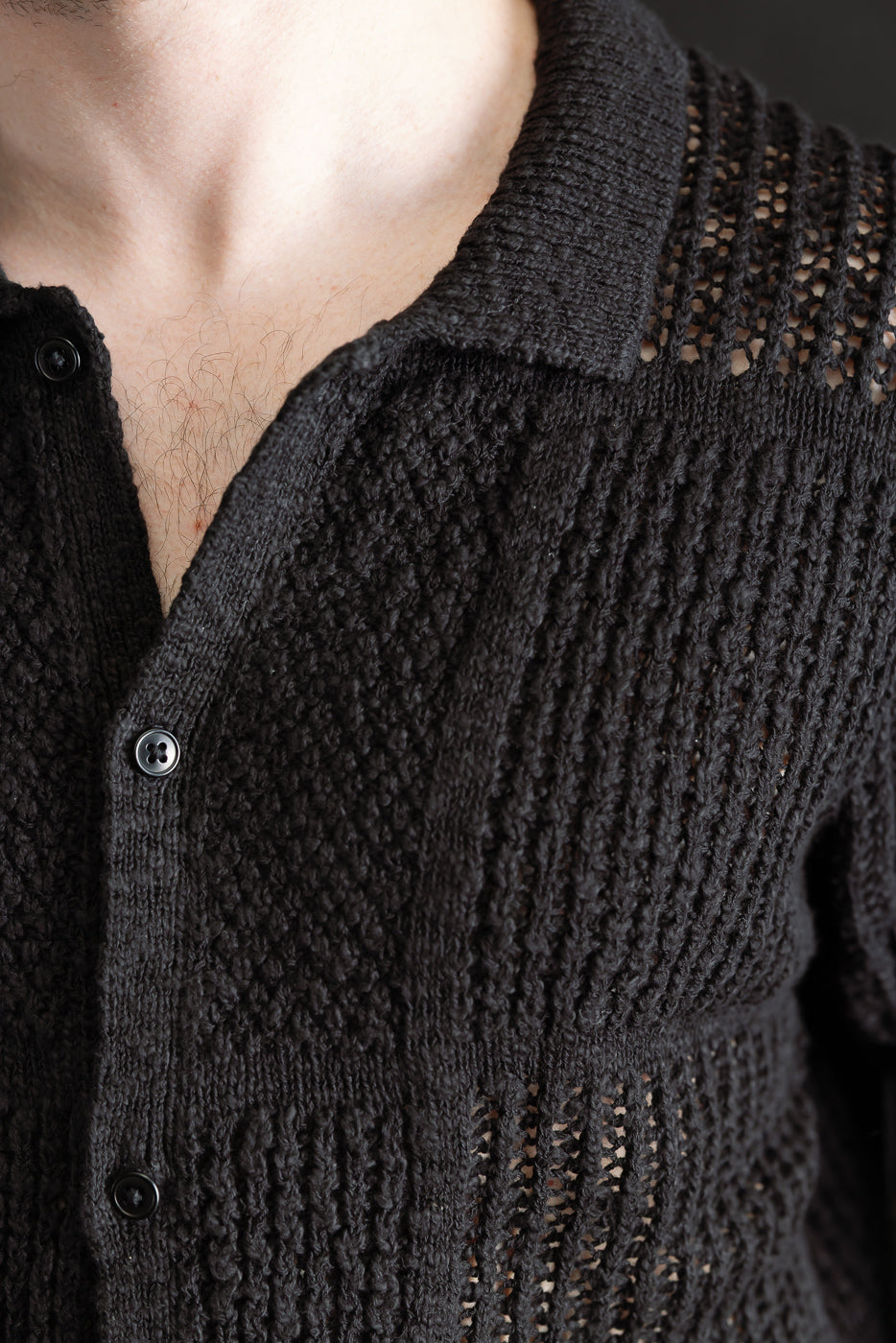 Close-up front of men's black knit short sleeve shirt on male model 5'9, 165lbs, showing textured knit, black buttons, open collar, and see-through upper fabric.