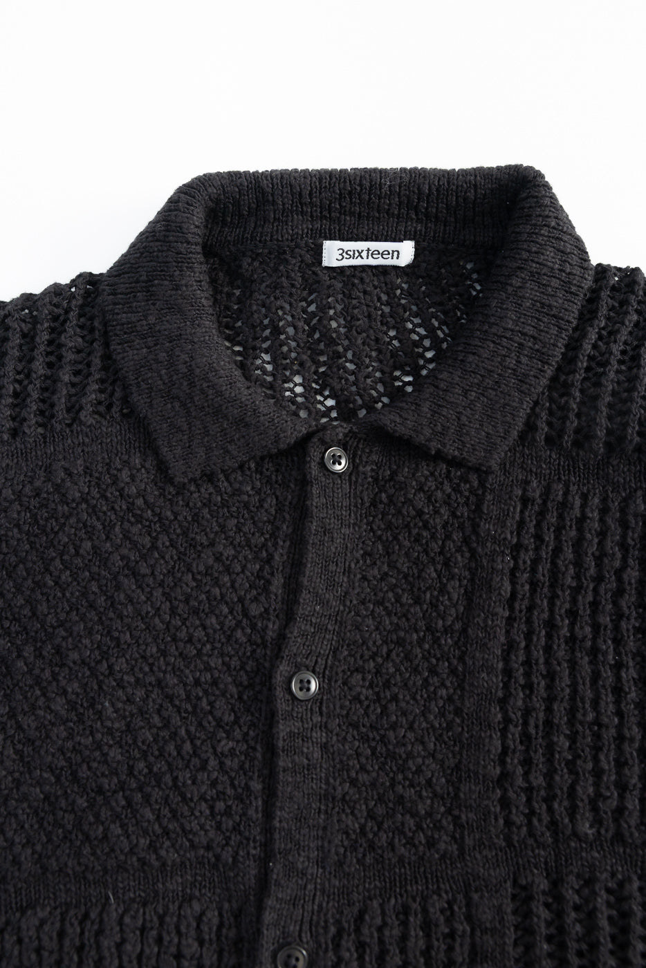 Close-up front view of black short sleeve knit shirt, showing textured fabric, buttons, and collar.