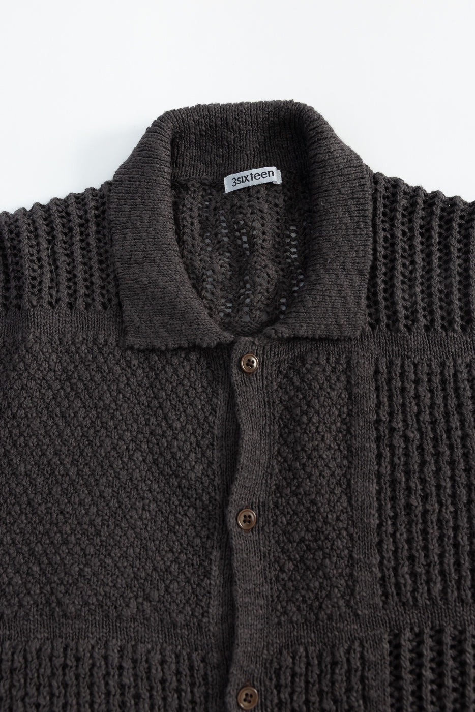 Close-up of front upper section of dark umber knit short sleeve shirt, showing textured fabric, button details, spread collar, and woven tag.