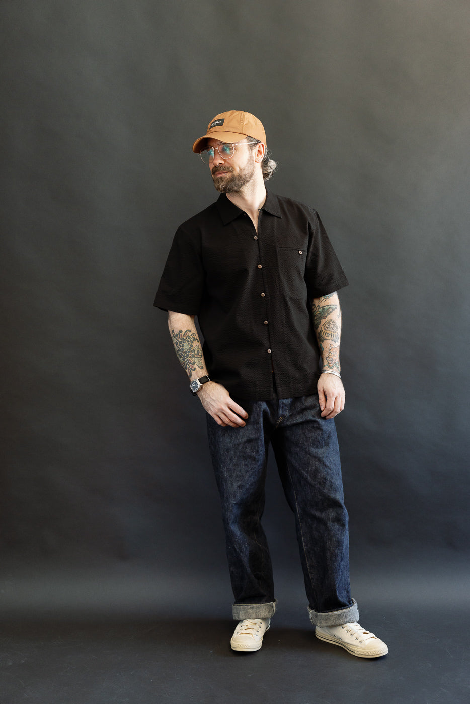 A casually dressed man models the Short Sleeve Loop Shirt - Black Seersucker, featuring a relaxed fit, open collar, and single chest pocket, paired with cuffed denim and canvas sneakers.