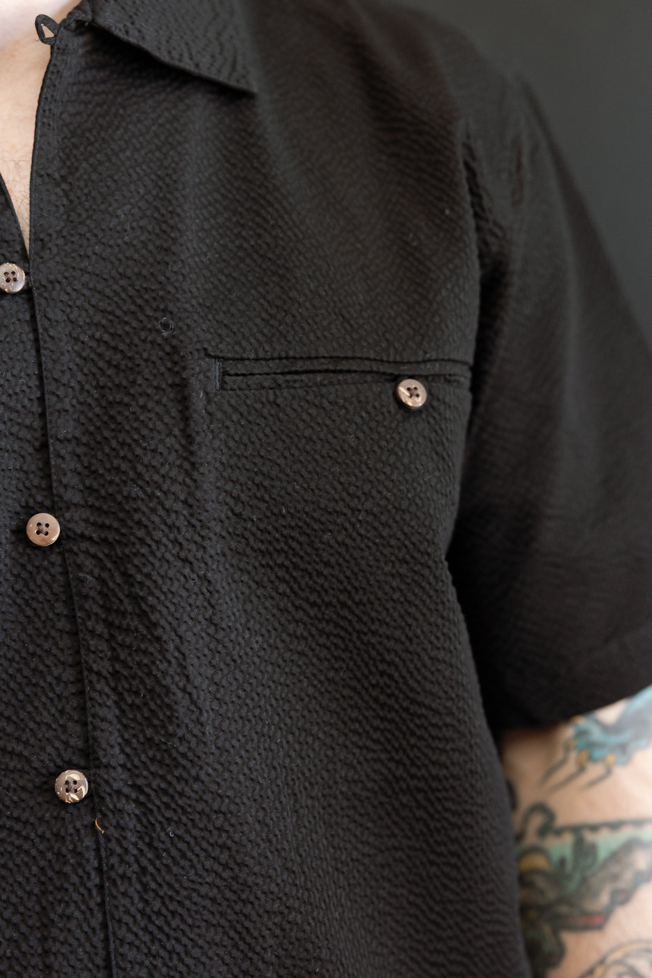 Close-up of the Short Sleeve Loop Shirt - Black Seersucker, highlighting its textured fabric, light-colored buttons, and single chest pocket, with a tattooed forearm partially visible.