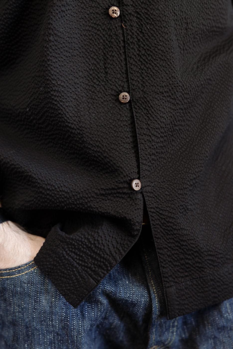 Close-up of the Short Sleeve Loop Shirt - Black Seersucker, showcasing its crinkled texture, small brown buttons, and a hand tucked into denim jeans, embodying Dant's premium menswear aesthetic.