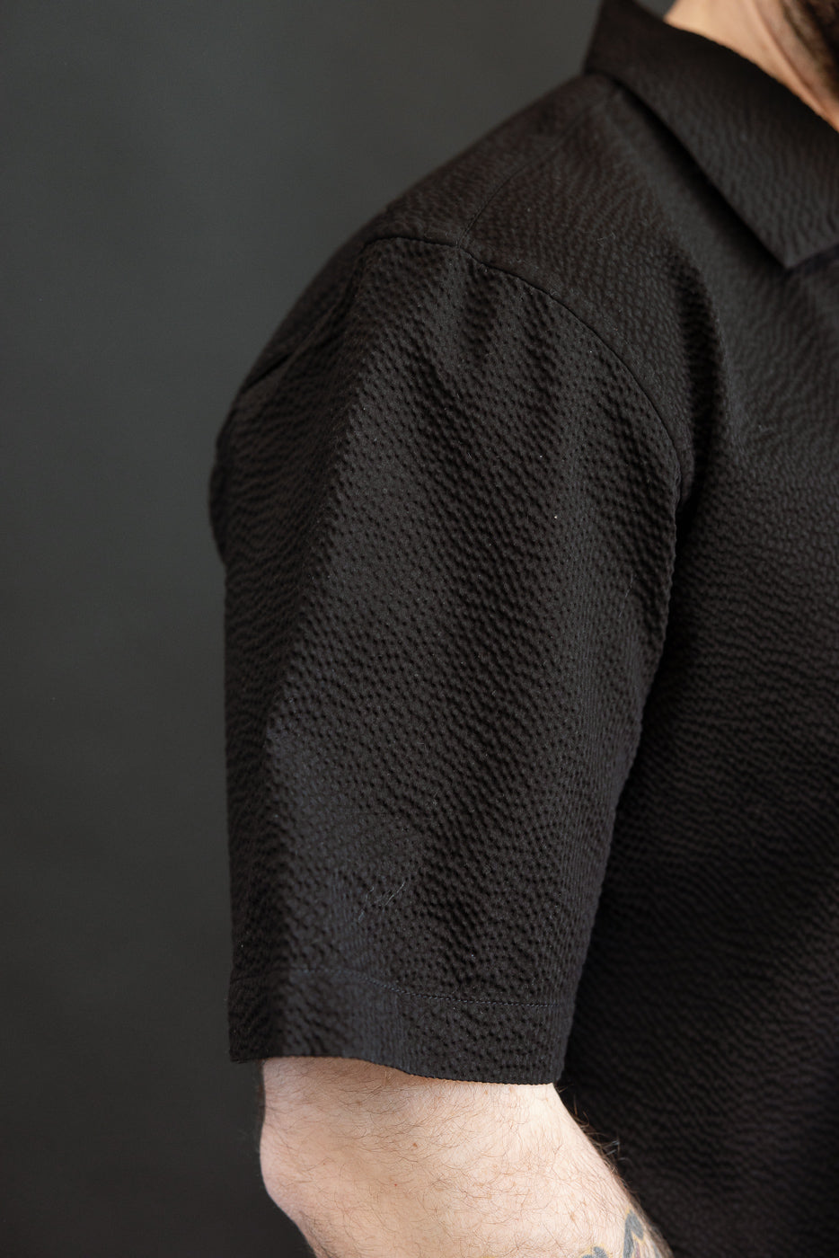 Close-up of a black, textured Short Sleeve Loop Shirt - Black Seersucker worn on an arm, showcasing the shirt's detailed fabric texture and short sleeves against a muted background.