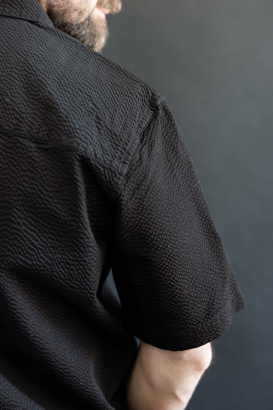 Person wearing Short Sleeve Loop Shirt - Black Seersucker, showcasing its textured fabric and short sleeves, with part of the forearm and beard visible, highlighting the shirt's distinctive utilitarian design.