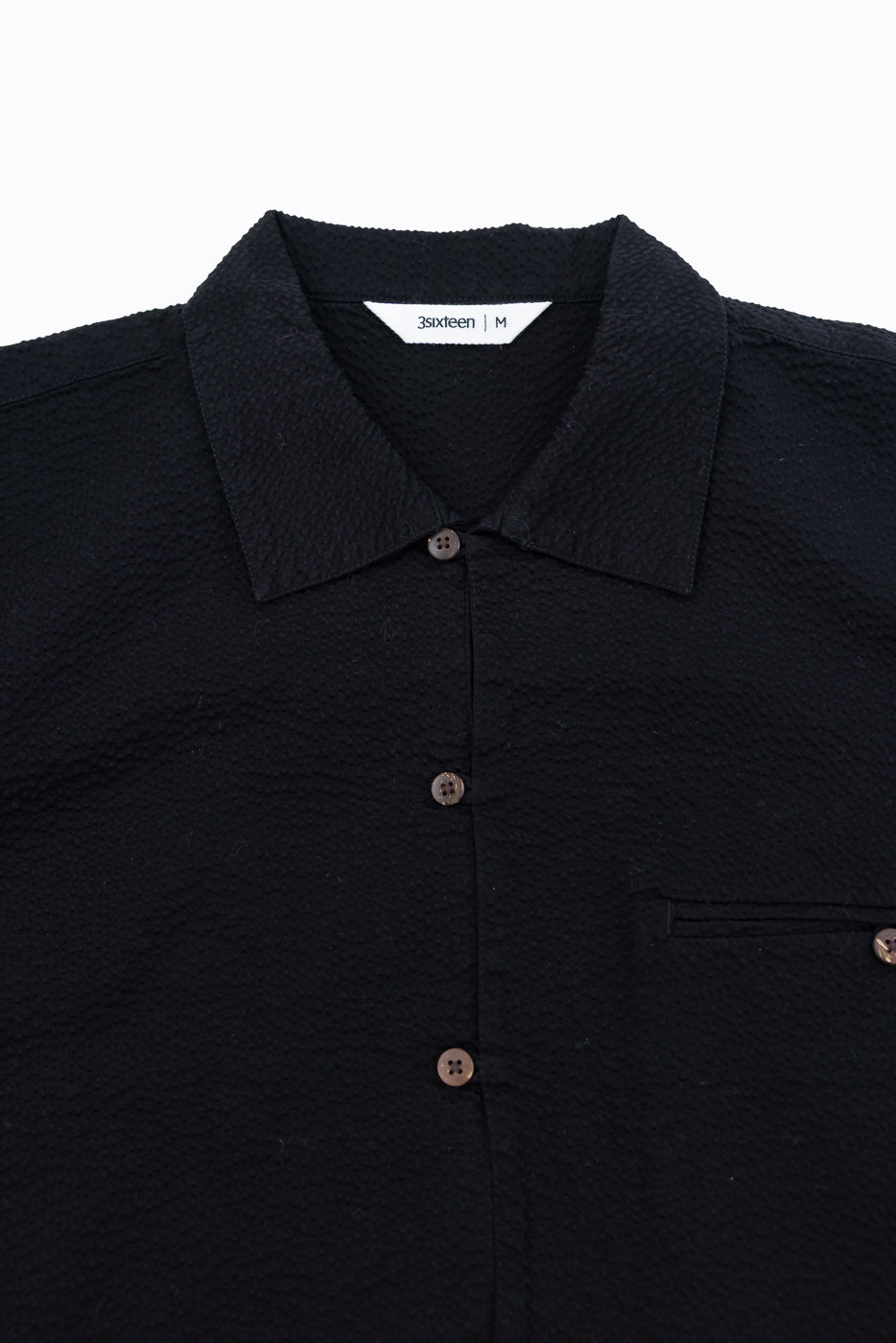 Short Sleeve Loop Shirt - Black Seersucker, featuring a textured short-sleeve design, pointed collar, dark brown buttons, and a chest pocket, laid flat, highlighting its refined craftsmanship and utilitarian style.