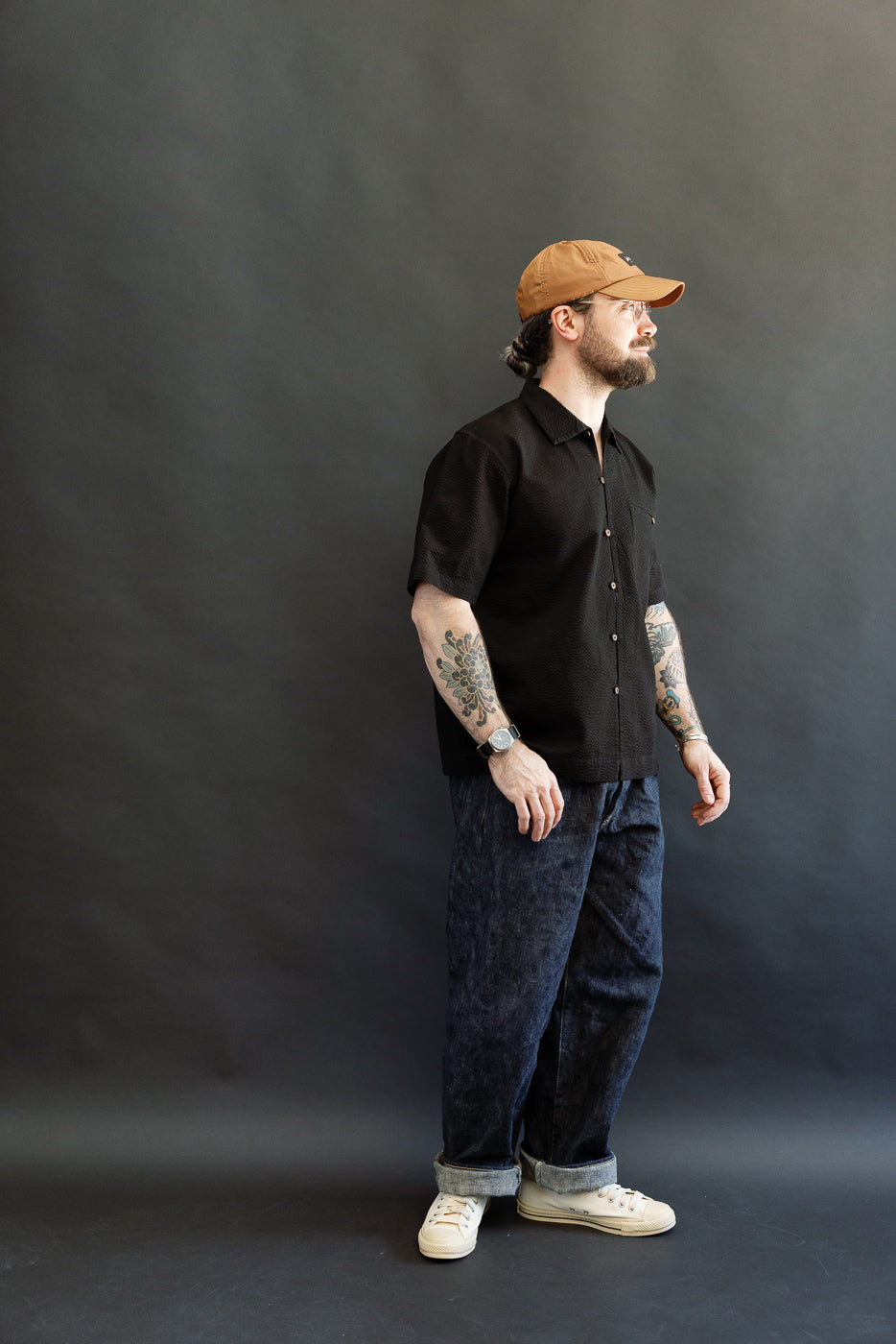 A Caucasian male model wearing the Short Sleeve Loop Shirt - Black Seersucker, featuring fabric loops and an open collar, paired with dark jeans and off-white sneakers, embodying a relaxed, heritage-inspired style.