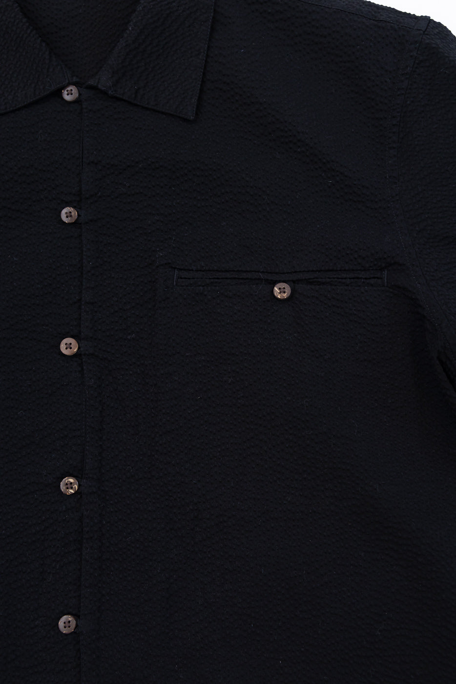 Close-up of the Short Sleeve Loop Shirt - Black Seersucker, featuring a textured cotton fabric, brown coconut shell buttons, an open collar, and a buttoned chest pocket, reflecting Dant's heritage-inspired style.