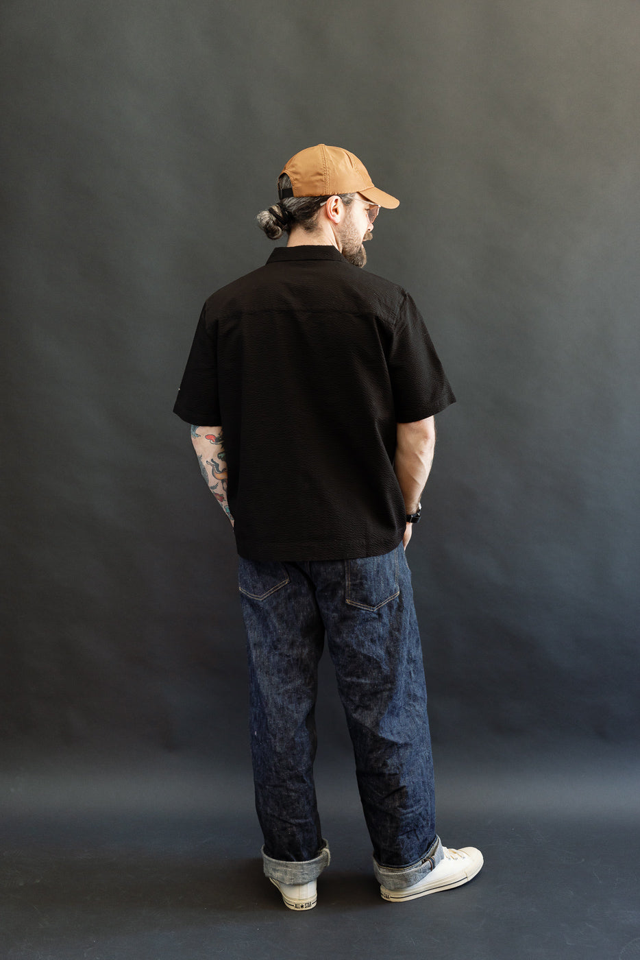 Man in Dant's Short Sleeve Loop Shirt - Black Seersucker, paired with dark denim jeans and white sneakers, showcasing casual contemporary style with visible tattoos and bun under a tan baseball cap.