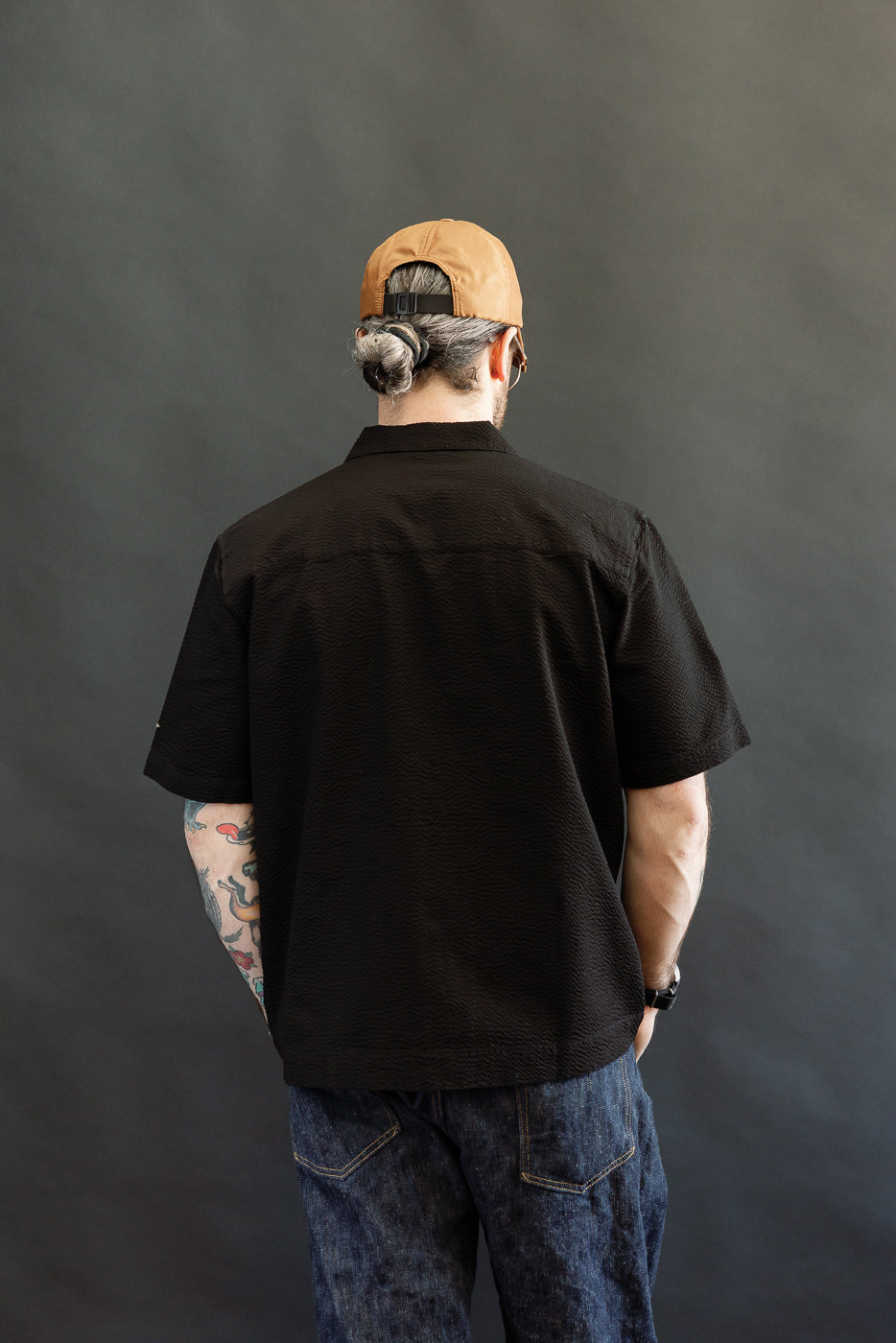 Person wearing Short Sleeve Loop Shirt - Black Seersucker, showcasing its textured fabric and relaxed fit, paired with classic blue jeans and a tan cap, standing casually with tattooed arm visible.