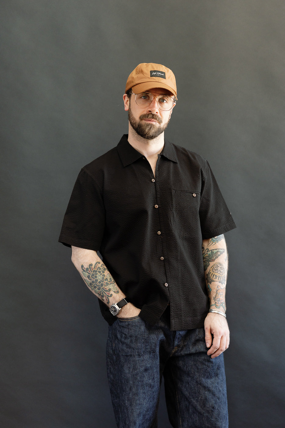 Model wearing the Short Sleeve Loop Shirt - Black Seersucker, showcasing colorful tattooed forearms. The shirt features a loop front placket, open collar, and coconut shell buttons, styled with dark denim jeans.