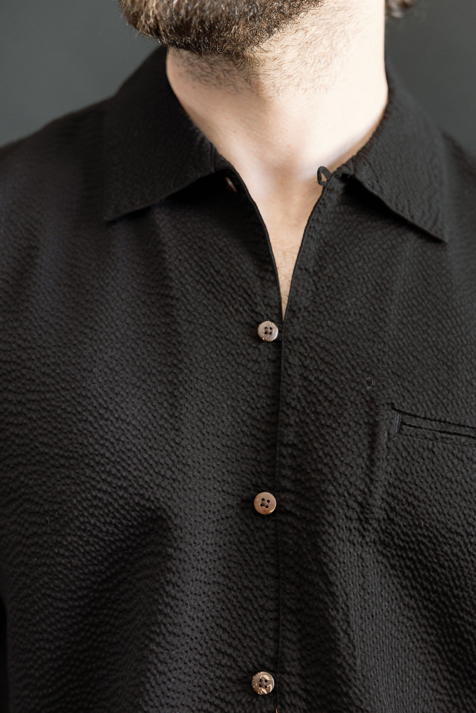Man wearing the Short Sleeve Loop Shirt - Black Seersucker with a textured surface, contrasting coconut shell buttons, open collar, and single chest pocket, showcasing Dant's heritage-inspired menswear collection.