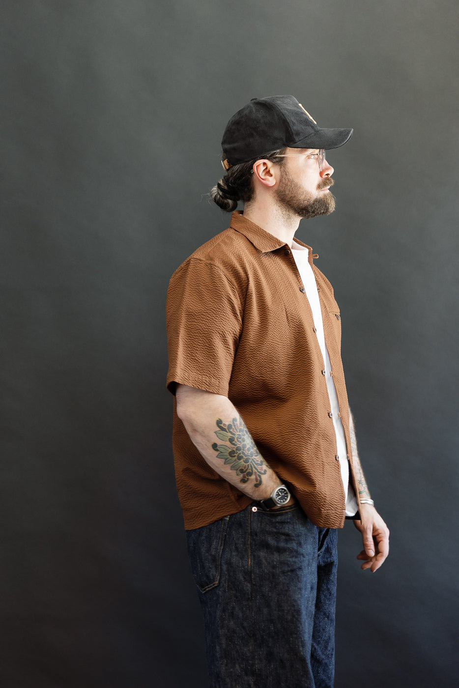 Man in profile wearing the Short Sleeve Loop Shirt - Cinnamon Seersucker with unique loop front placket, open collar, single chest pocket, and coconut shell buttons, styled over a white T-shirt.