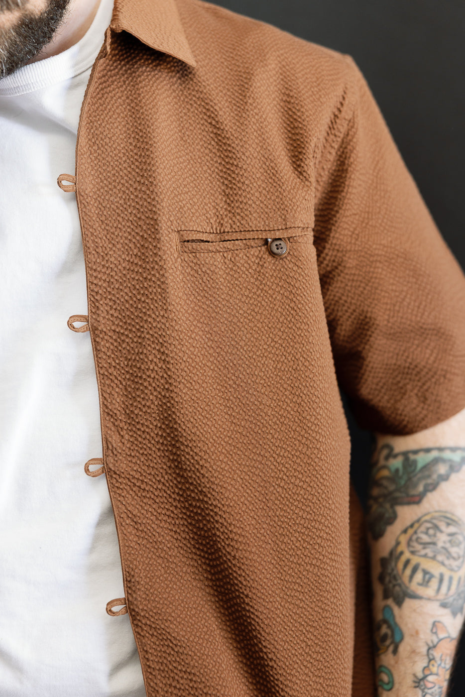 A close-up of the Short Sleeve Loop Shirt - Cinnamon Seersucker, showcasing its textured brown seersucker fabric, unique loop placket, and chest pocket, worn over a white T-shirt.