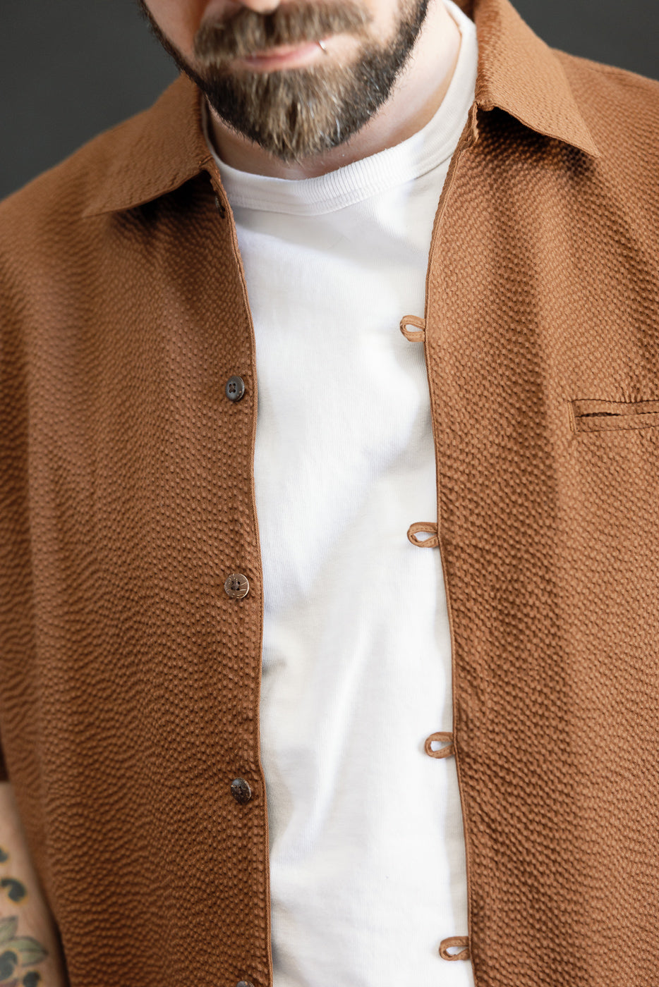 The Short Sleeve Loop Shirt - Cinnamon Seersucker is showcased with its open collar and textured fabric, featuring unique fabric loops and coconut shell buttons, layered over a white tee.