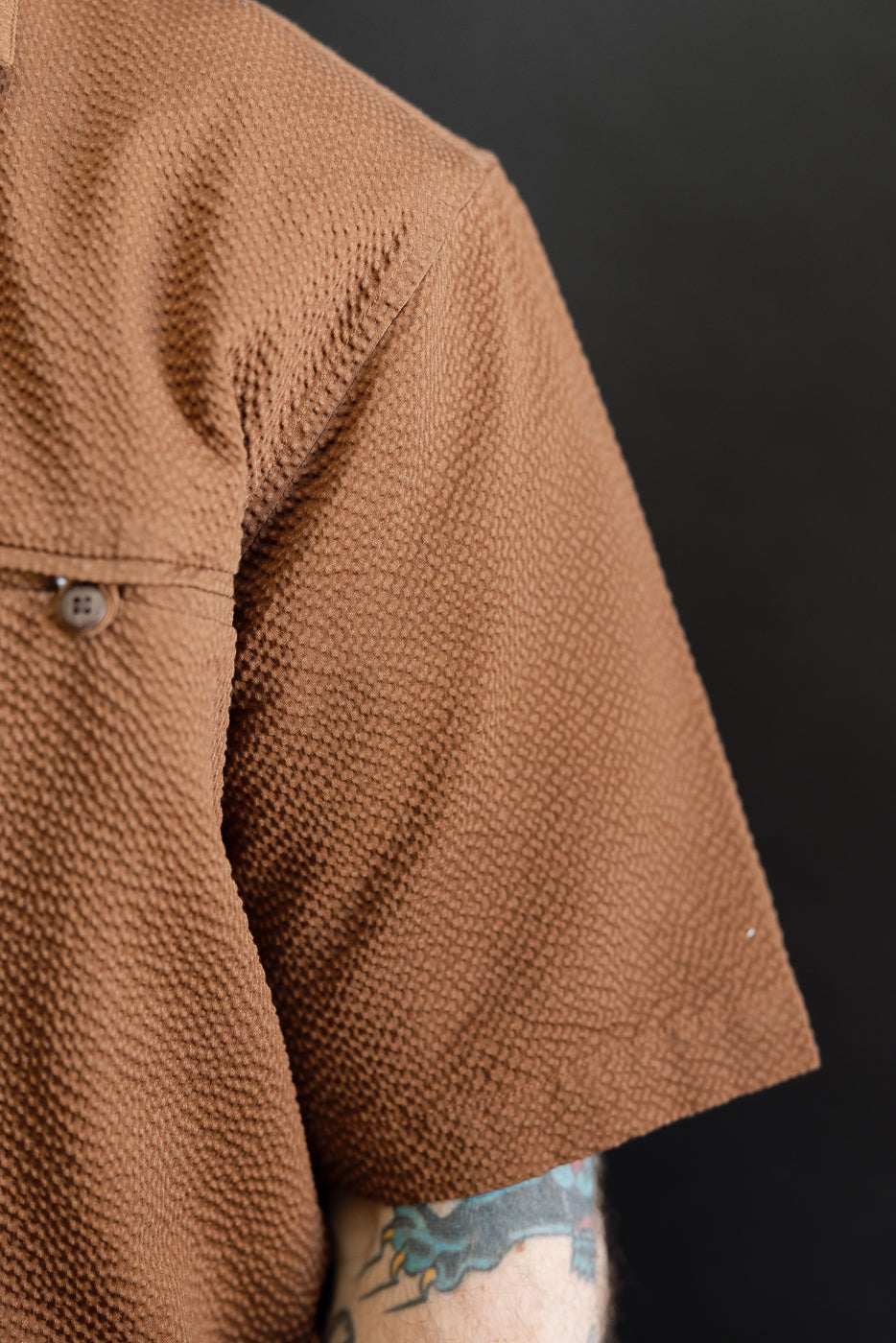 Close-up of the Short Sleeve Loop Shirt - Cinnamon Seersucker, highlighting its textured waffle-like knit, flap pocket with button on the back yoke, and the hint of the wearer's tattooed arm.