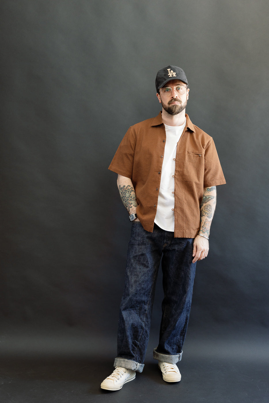 Model wearing the Short Sleeve Loop Shirt - Cinnamon Seersucker, featuring an open collar and loop placket, styled over a white tee. Paired with rolled-up denim and sneakers, showcasing casual sophistication.