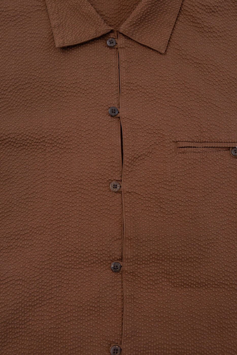 Close-up of the Short Sleeve Loop Shirt - Cinnamon Seersucker, featuring a textured seersucker fabric, open collar, fabric loop placket, matching brown coconut shell buttons, and a single chest pocket on the right.
