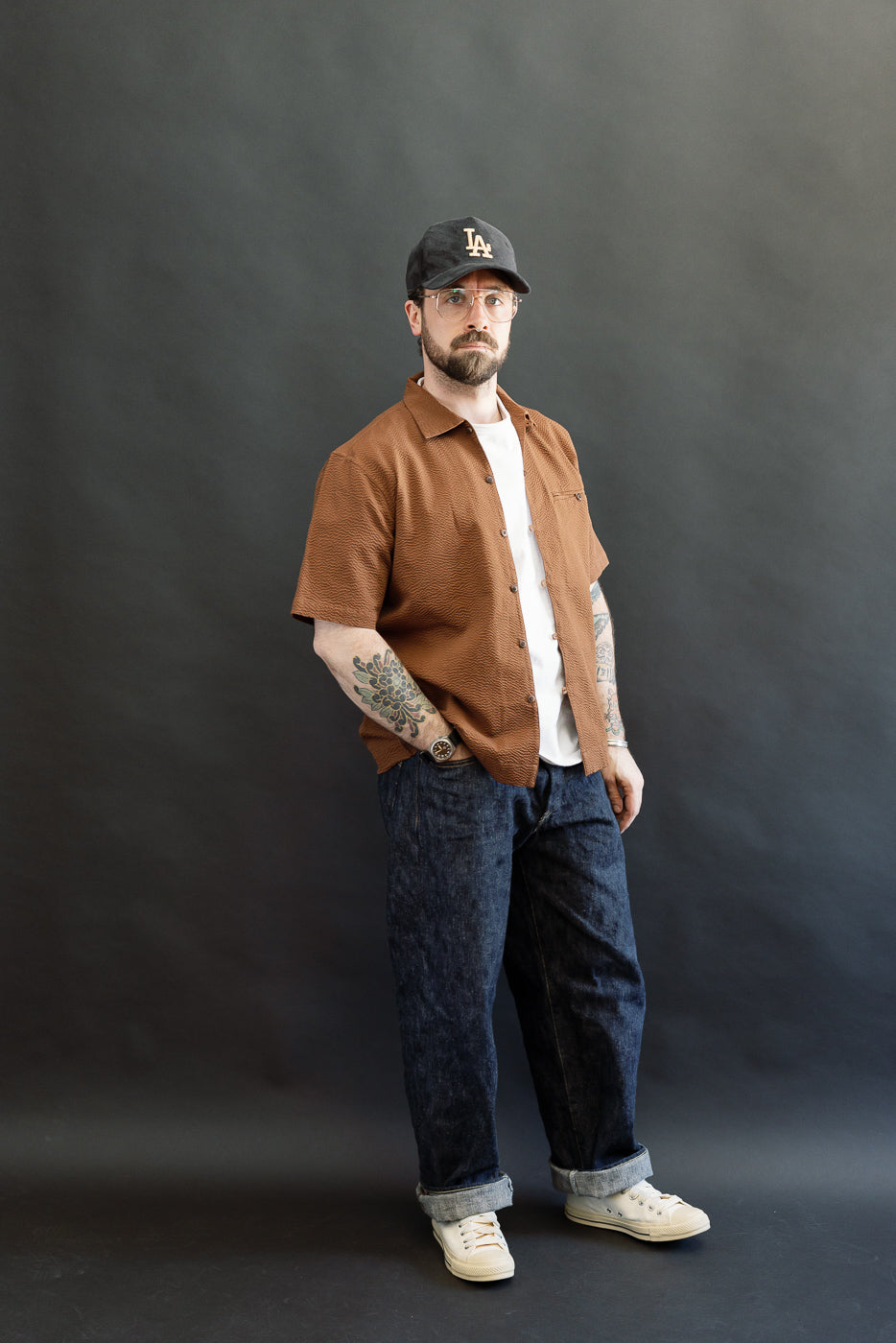 Man wearing the Short Sleeve Loop Shirt - Cinnamon Seersucker, featuring a loop front placket and open collar, styled over a white T-shirt with rolled-up dark denim jeans and off-white sneakers.