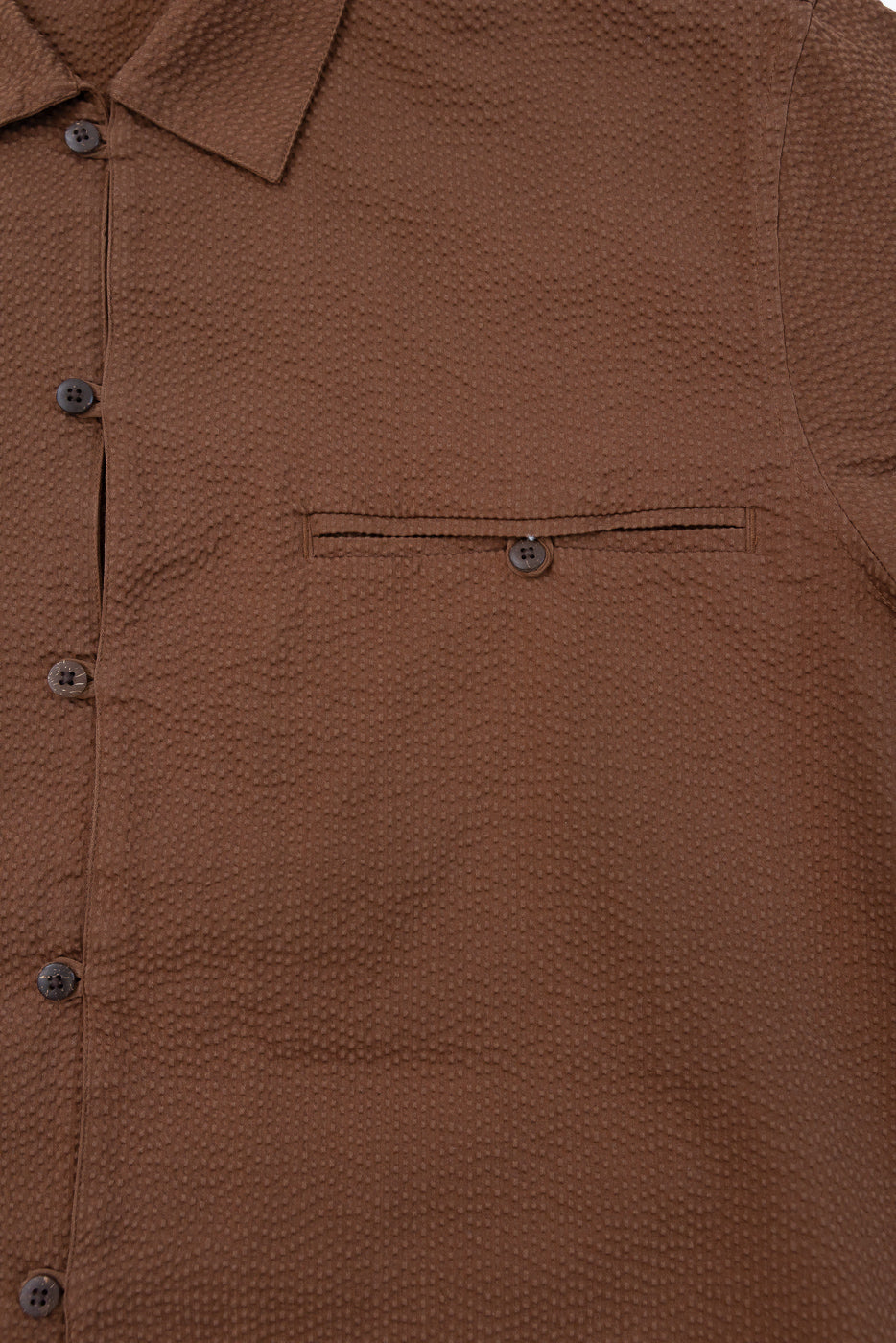 Close-up of the Short Sleeve Loop Shirt - Cinnamon Seersucker, showcasing its textured brown fabric, distinctive loop front placket, coconut shell buttons, open collar, and single buttoned chest pocket.