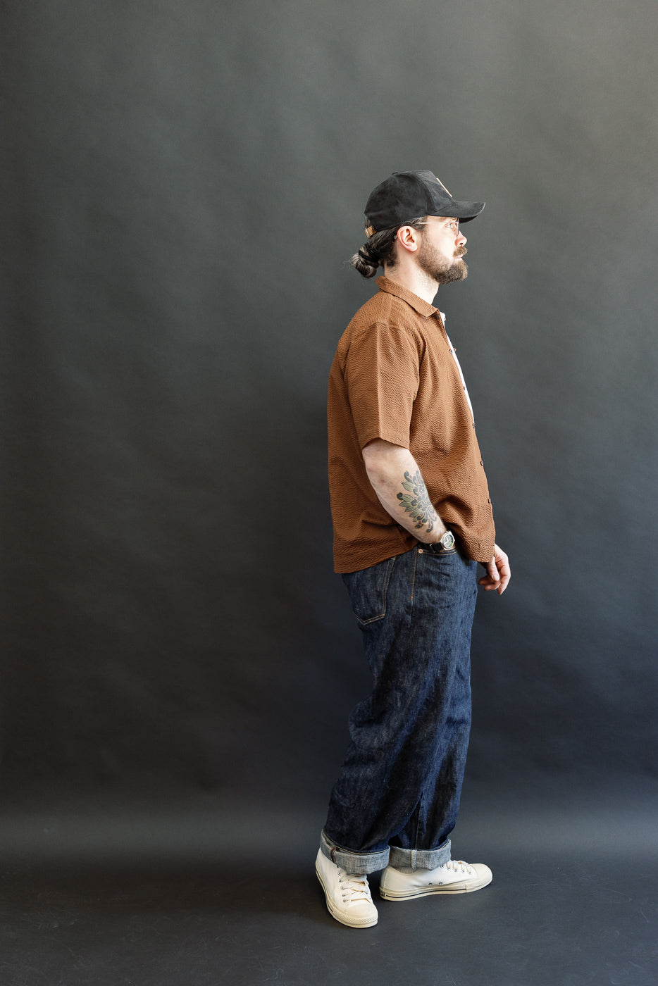 A man models the 3sixteen Short Sleeve Loop Shirt in cinnamon seersucker, showcasing its textured fabric and loop front placket. Styled with dark blue jeans and white sneakers, he exudes casual elegance.