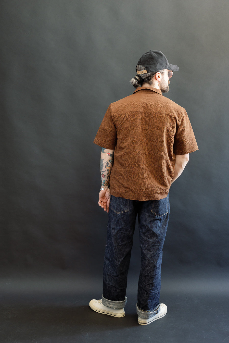 Man in Short Sleeve Loop Shirt - Cinnamon Seersucker, showcasing its unique loop front placket and textured design, paired with dark denim jeans and off-white sneakers, embodying casual elegance.
