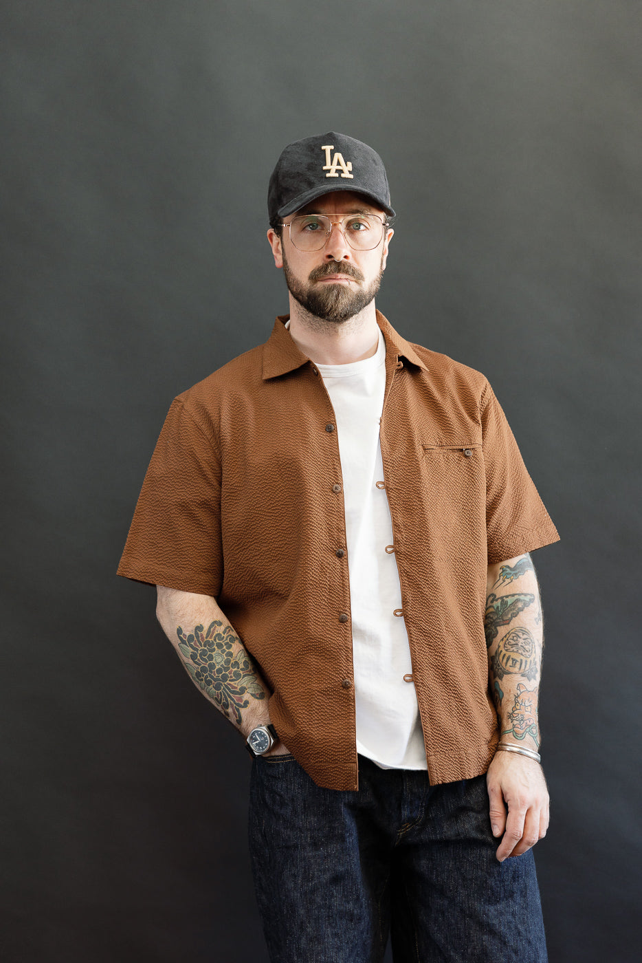 Man wearing Short Sleeve Loop Shirt - Cinnamon Seersucker, featuring a unique loop front placket, open collar, and single chest pocket, layered over a white T-shirt, paired with dark jeans and accessories.