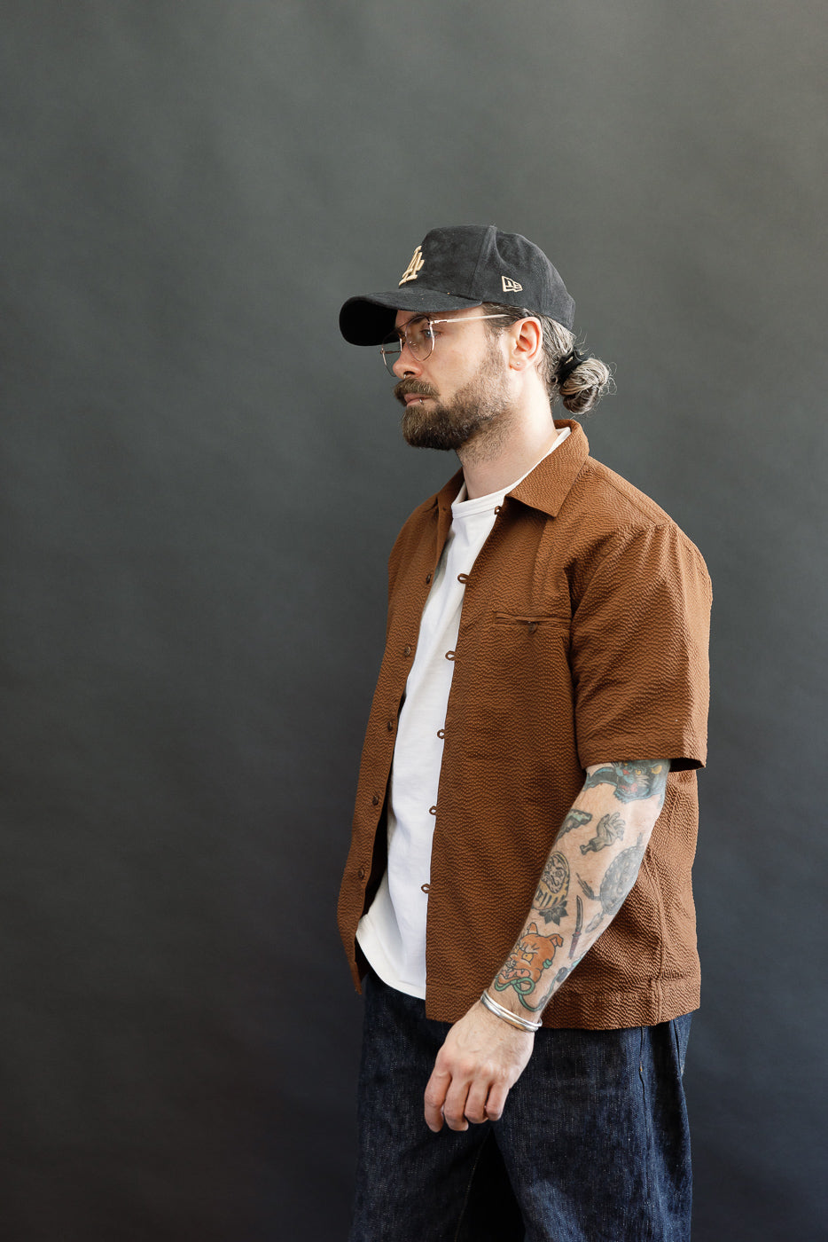 Man wearing the Short Sleeve Loop Shirt in cinnamon seersucker, featuring a loop front placket, open collar, and coconut shell buttons, styled with a white T-shirt and dark jeans in a casual look.