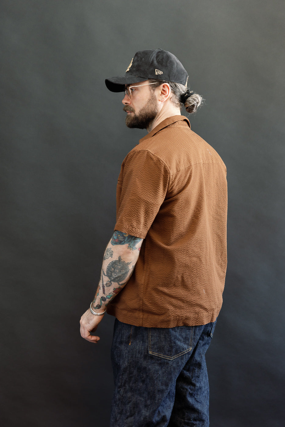 Man in three-quarter profile wearing the Short Sleeve Loop Shirt - Cinnamon Seersucker with fabric loops, textured brown fabric, open collar, and coconut shell buttons, styled with dark denim jeans and a black baseball cap.