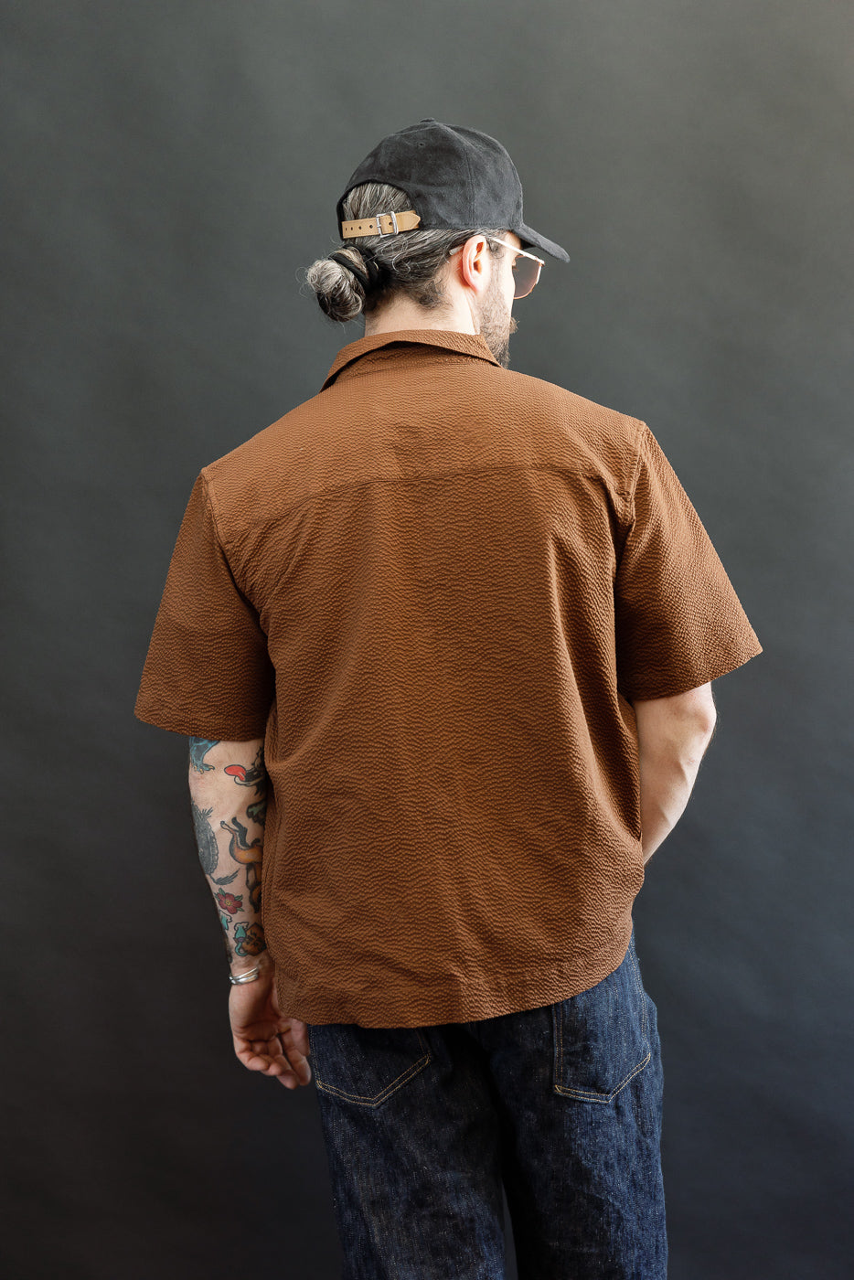 A man models the Short Sleeve Loop Shirt - Cinnamon Seersucker, showcasing its distinctive loop front placket and open collar, complemented by dark blue jeans and a backwards black cap.