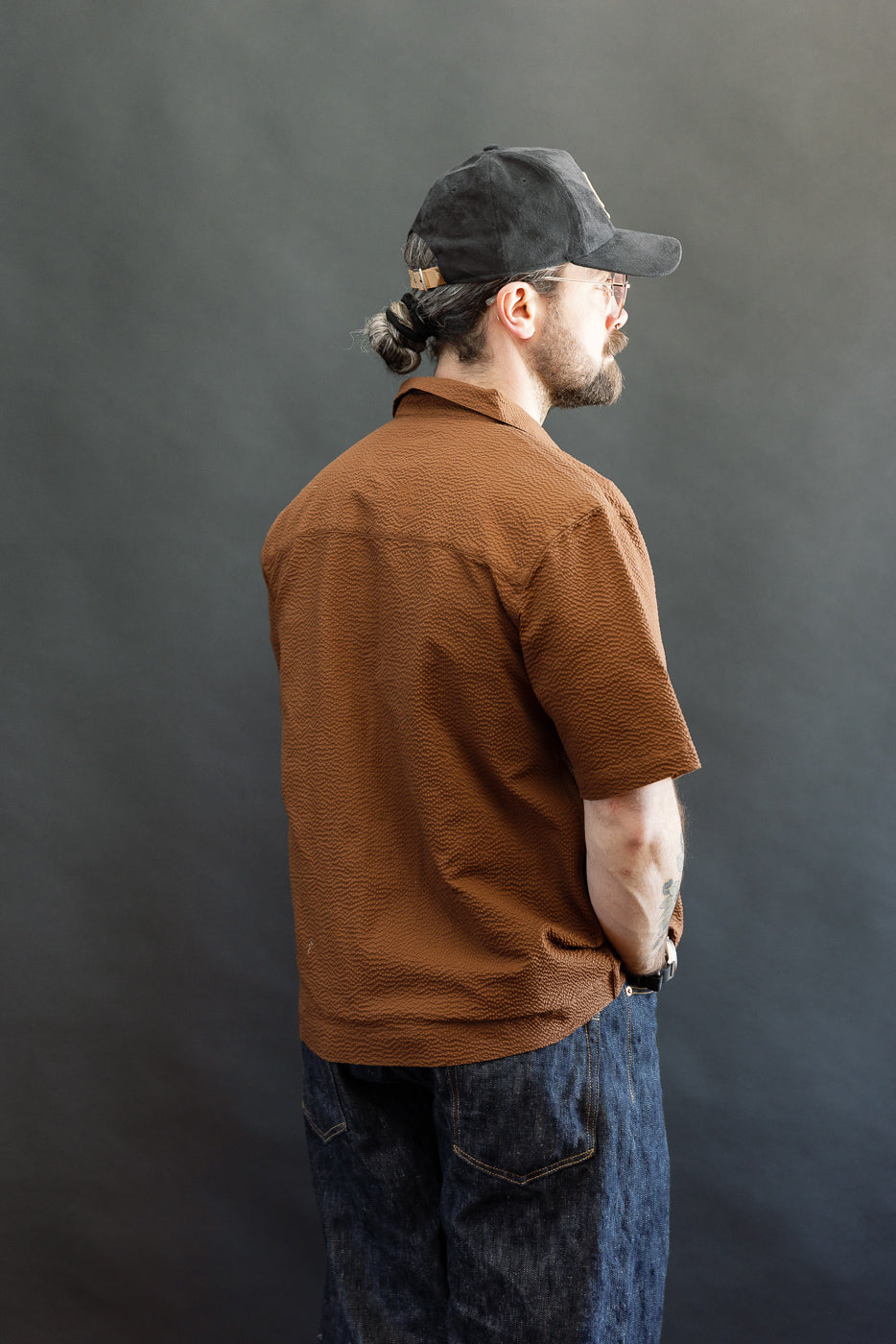 A man models the Short Sleeve Loop Shirt - Cinnamon Seersucker, showcasing its textured fabric, loop placket, and open collar style, complemented by casual loose dark jeans and a black baseball cap.