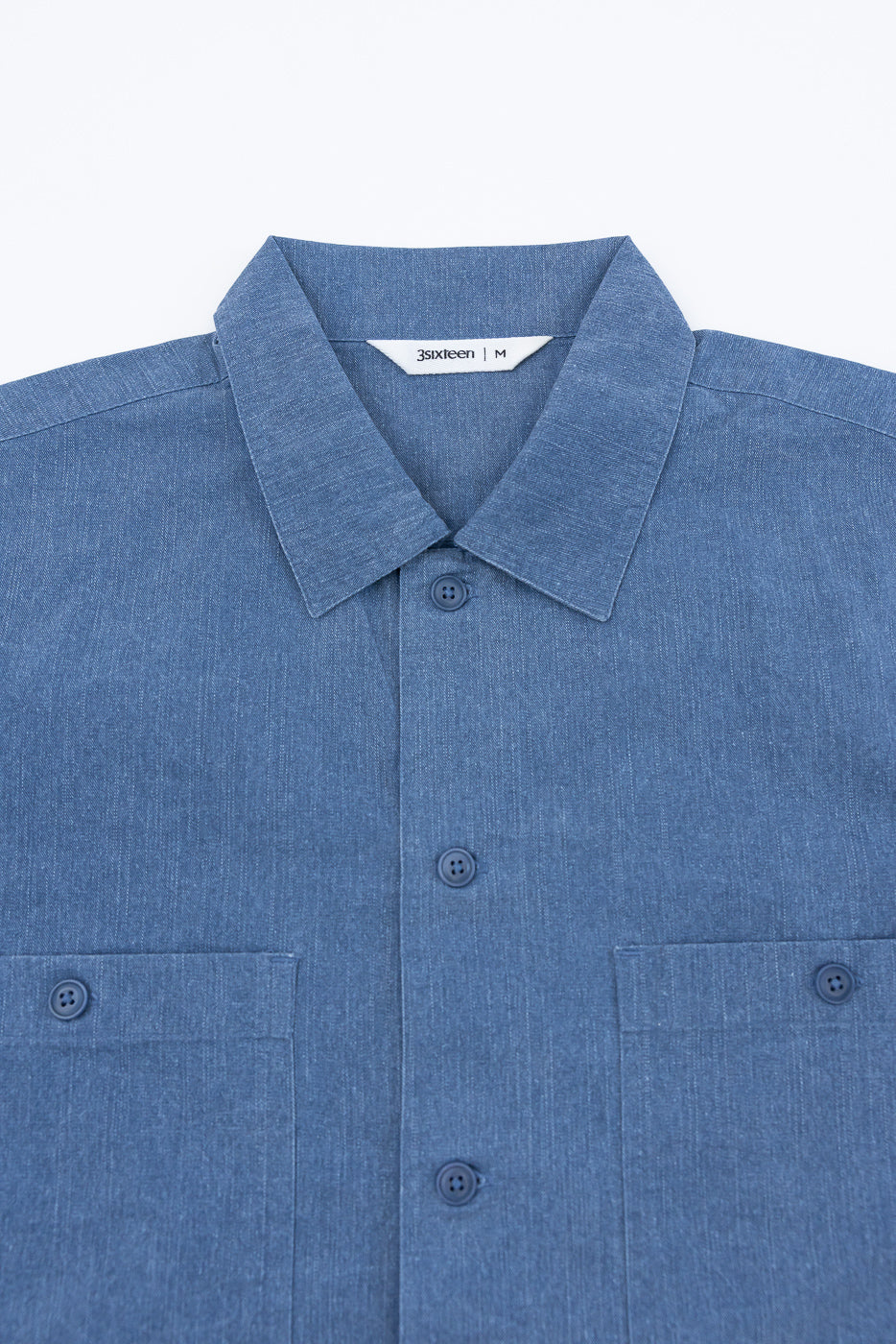 Blue, long-sleeve button-up shirt with a classic point collar, matching buttons, and dual chest pockets. Made from mid-weight chambray, labeled 3sixteen | M, reflecting modern workwear style.