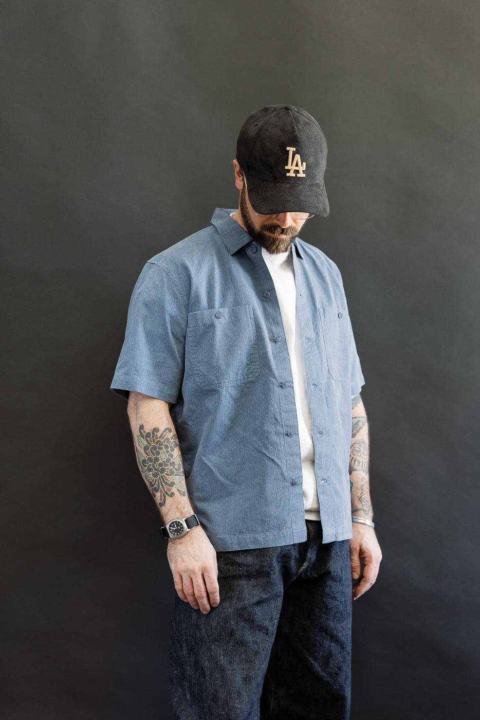 Man wearing a 3sixteen Short Sleeve Work Shirt - Blue Pigment Dye, styled open over a white T-shirt, showcasing dual chest pockets and Corozo buttons, paired with dark denim jeans and clear-rimmed glasses.